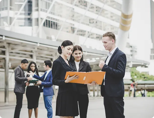 Three business professionals standing outdoors discussing an orange folder.