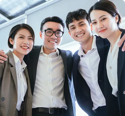 Four young business professionals in suits smiling and posing closely together indoors.