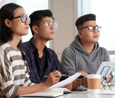 Three young adults wearing glasses attentively listening and taking notes in a classroom.