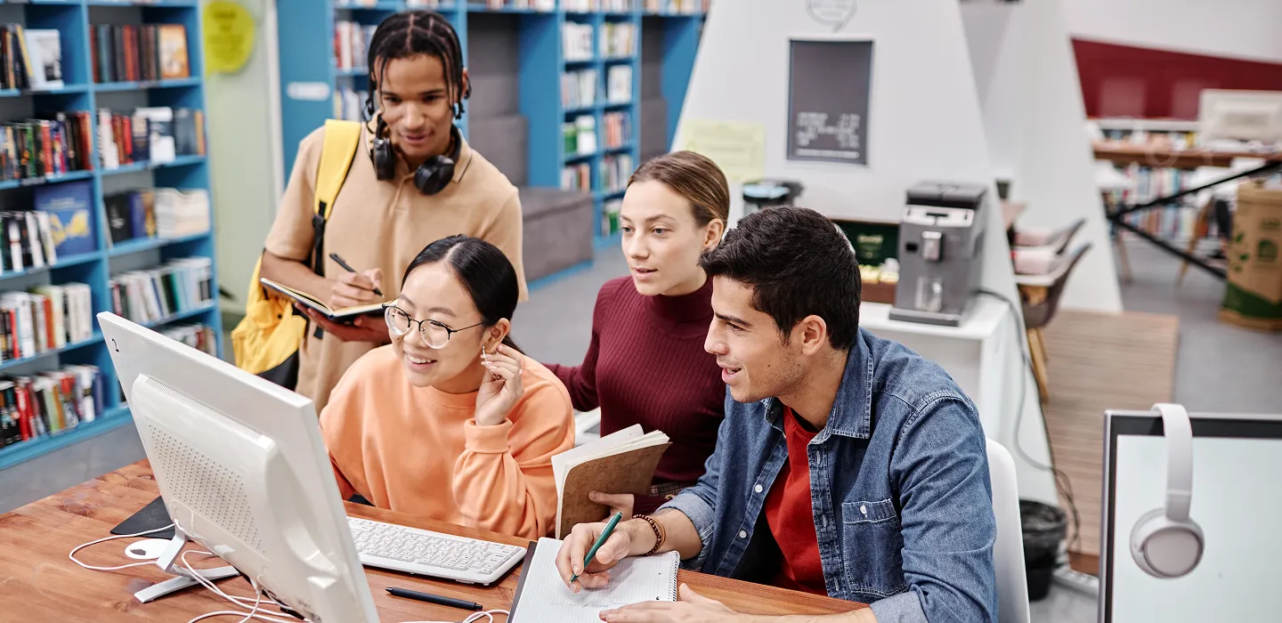 Four diverse students studying together in a library, working on a computer and taking notes.