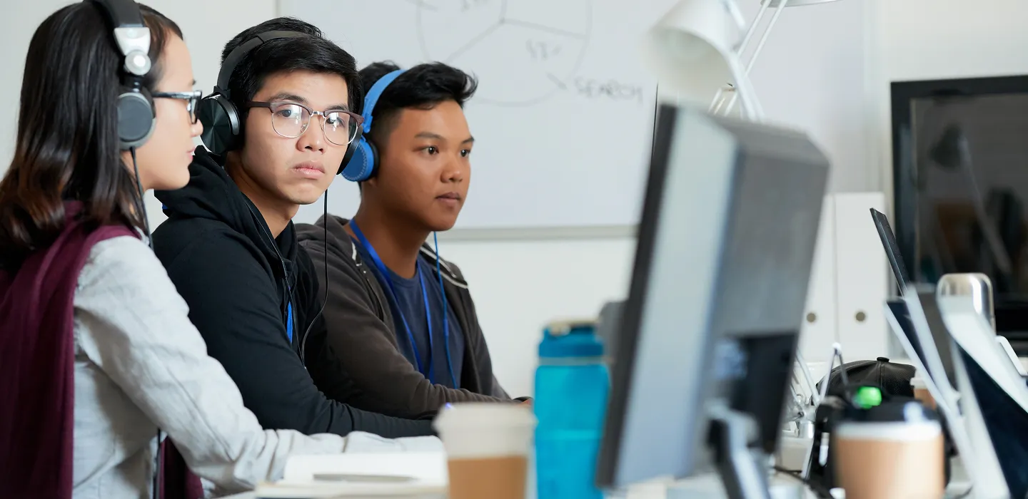 Three young people wearing headphones working on computers in a classroom or office setting.