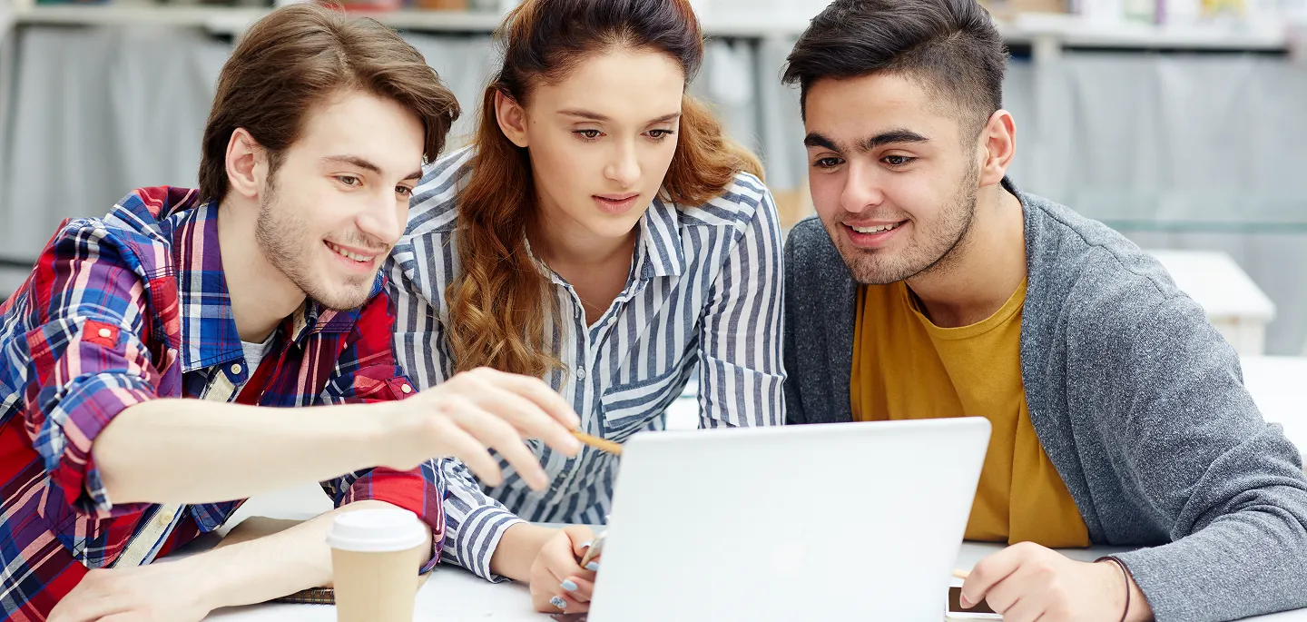 Three young adults looking at a laptop screen together, one pointing with a pencil.