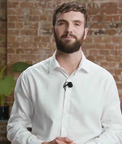 Man with beard wearing white shirt and lapel microphone in front of a brick wall.