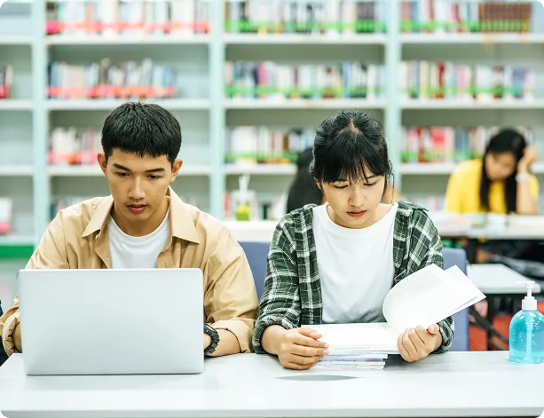 Two students studying at a library table; one uses a laptop, the other reads a book.