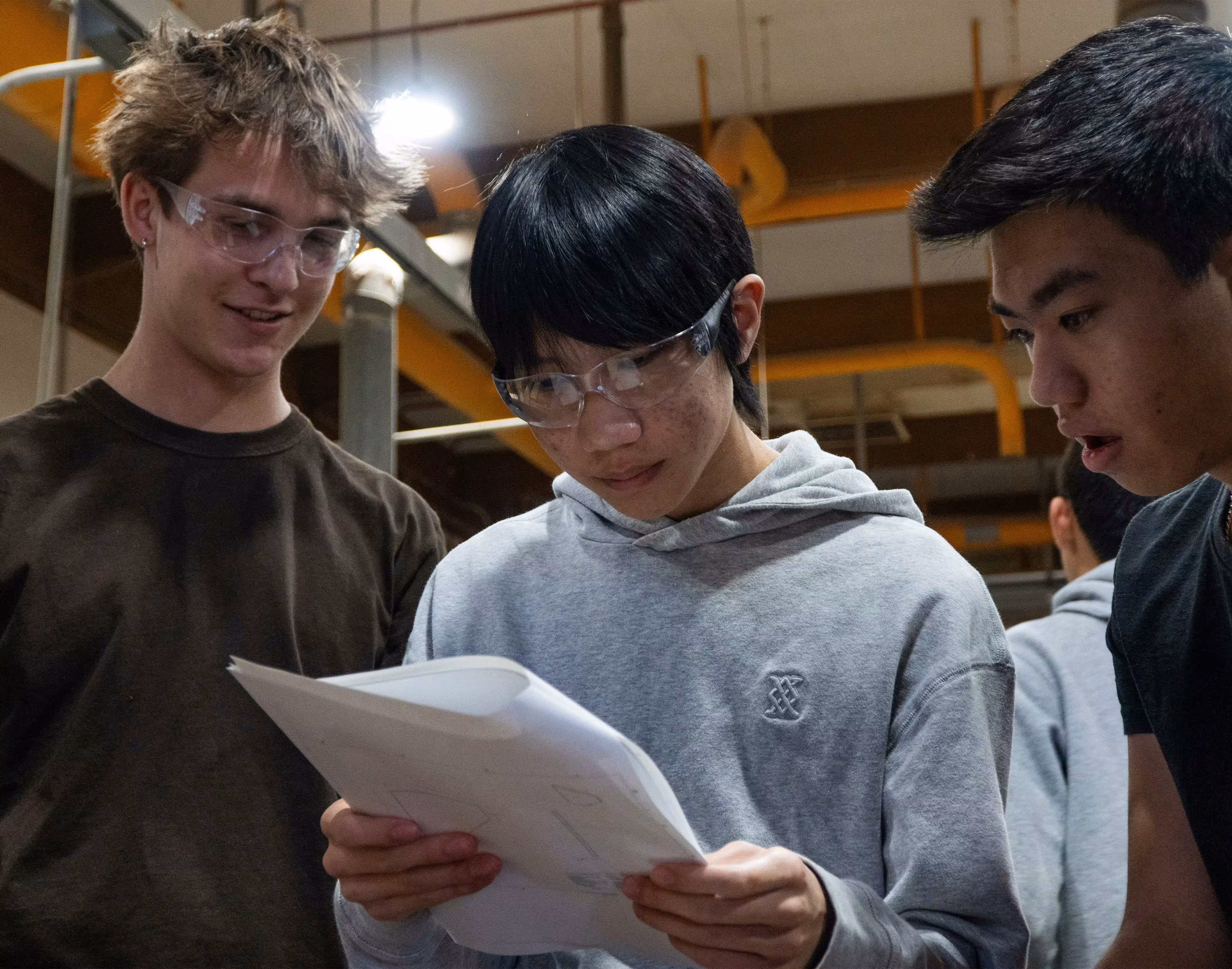 Three young men wearing safety glasses reviewing papers together in an industrial setting.