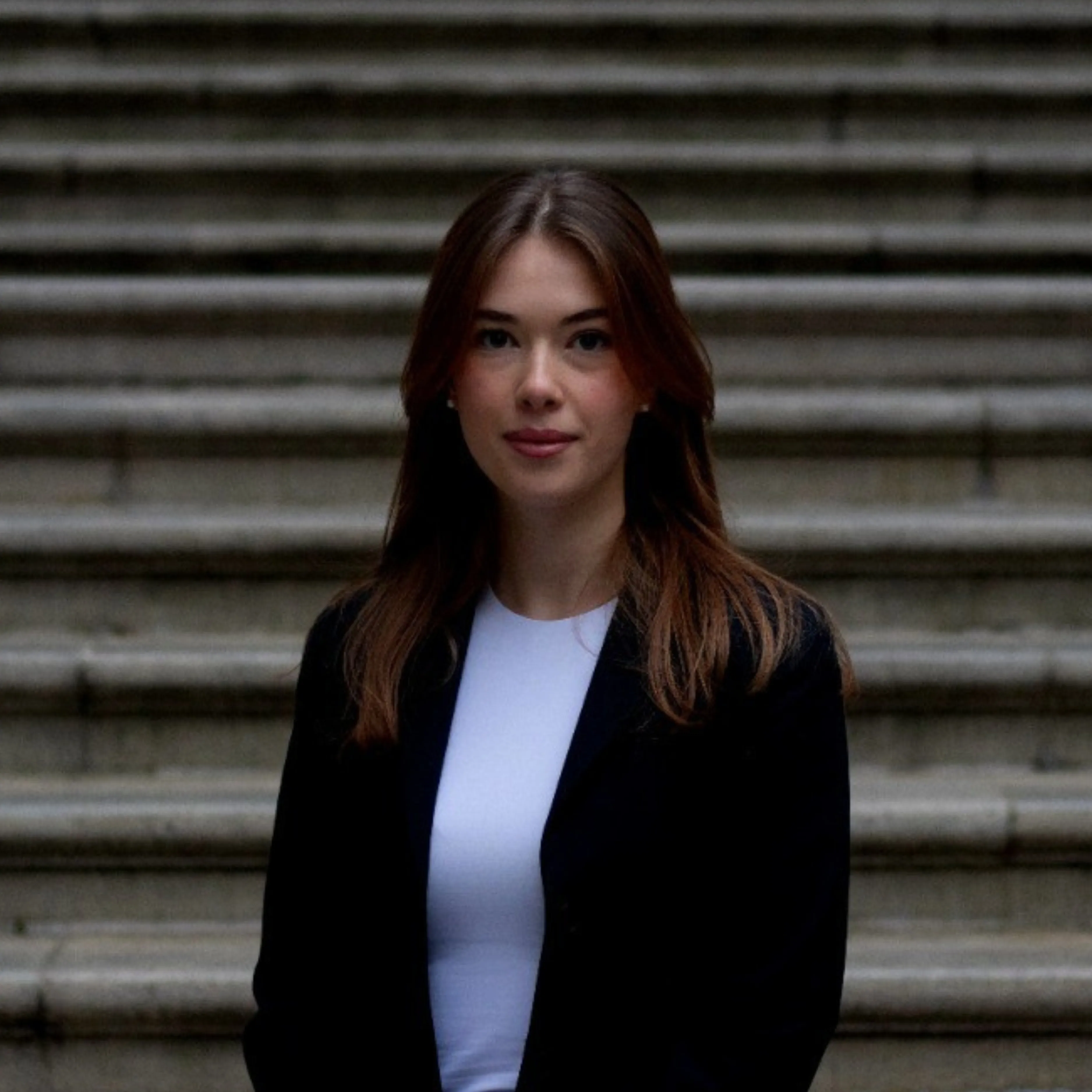 Young woman with brown hair in a black blazer and white top sitting in front of stone steps.