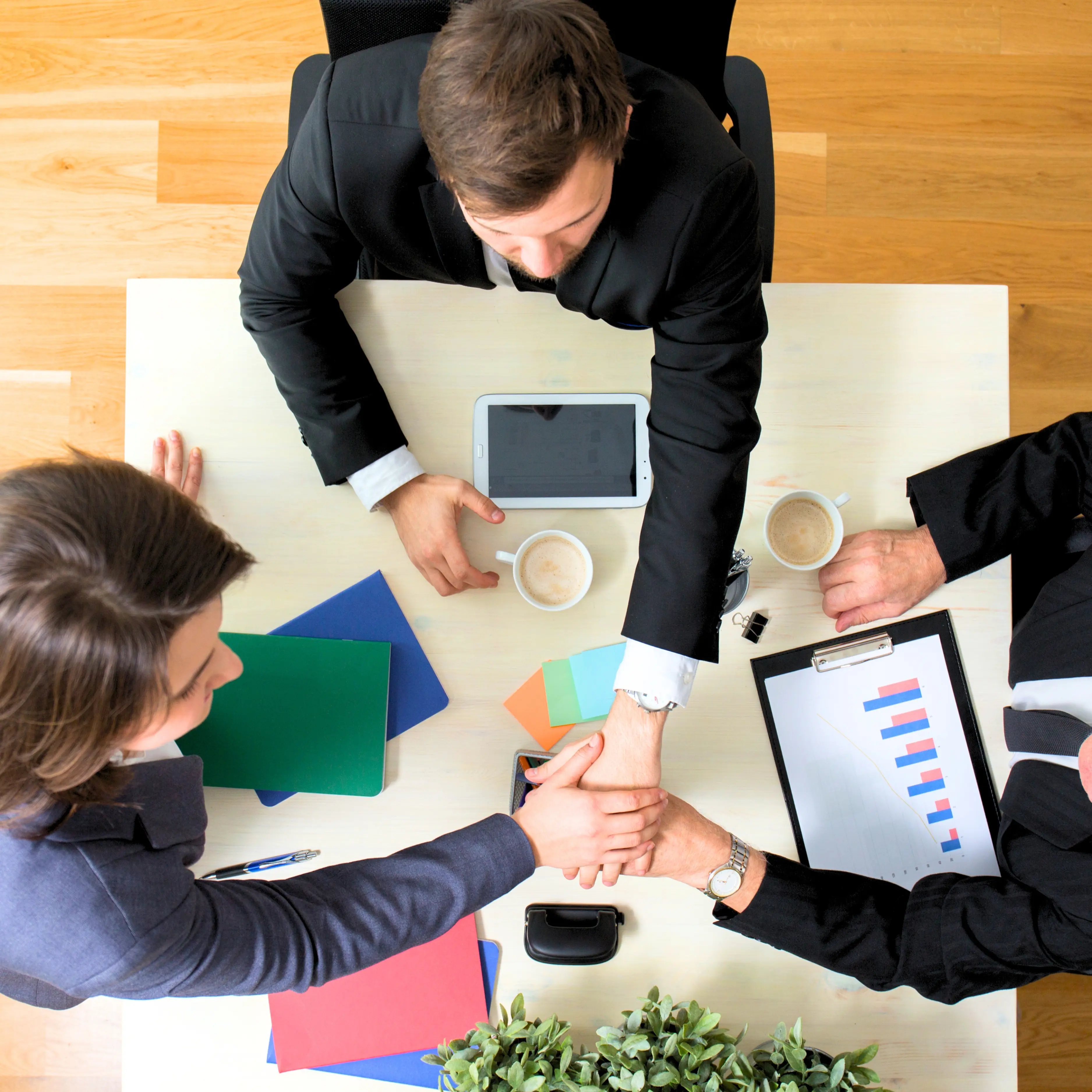 Three businesspeople in suits joining hands over a table with coffee and documents.