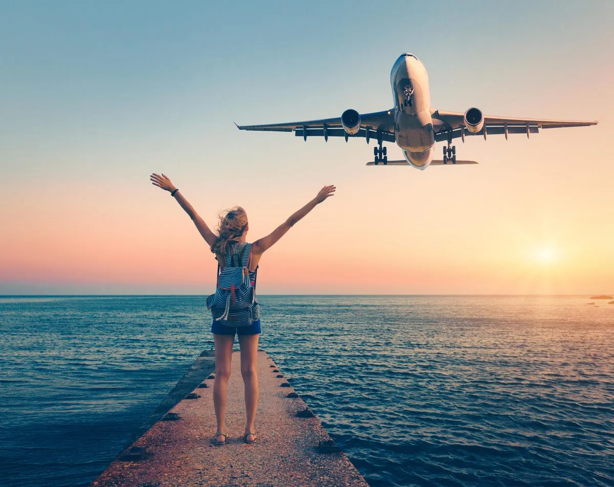 Woman with backpack on pier raises arms as airplane flies low over ocean at sunset.