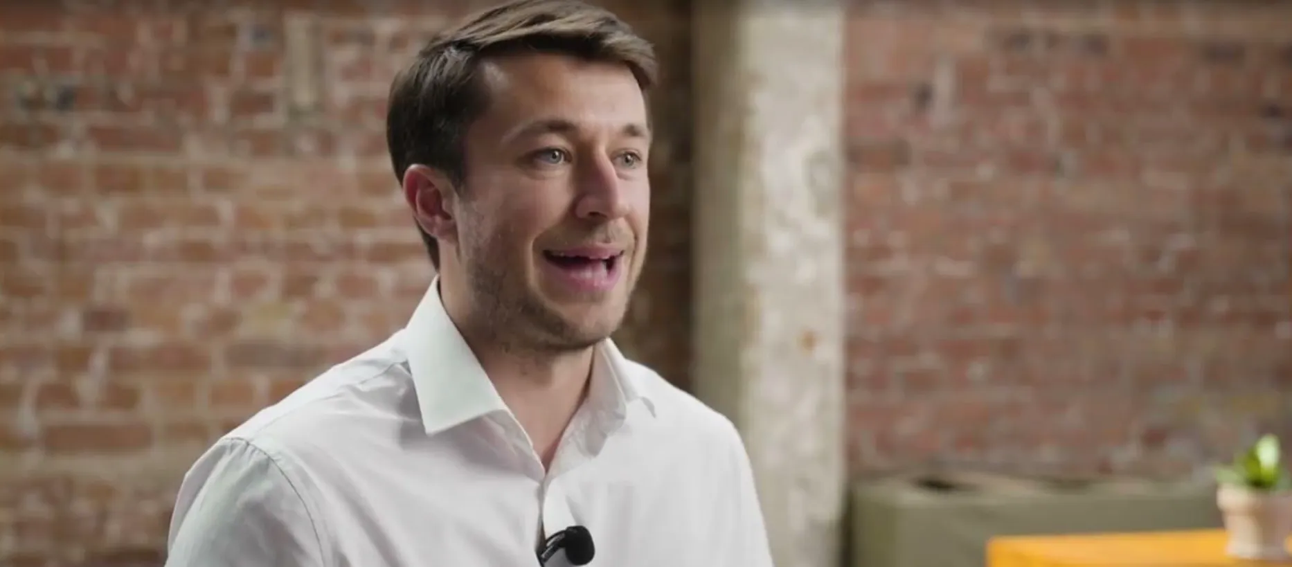 Man with short brown hair and white shirt speaking in front of a brick wall background.