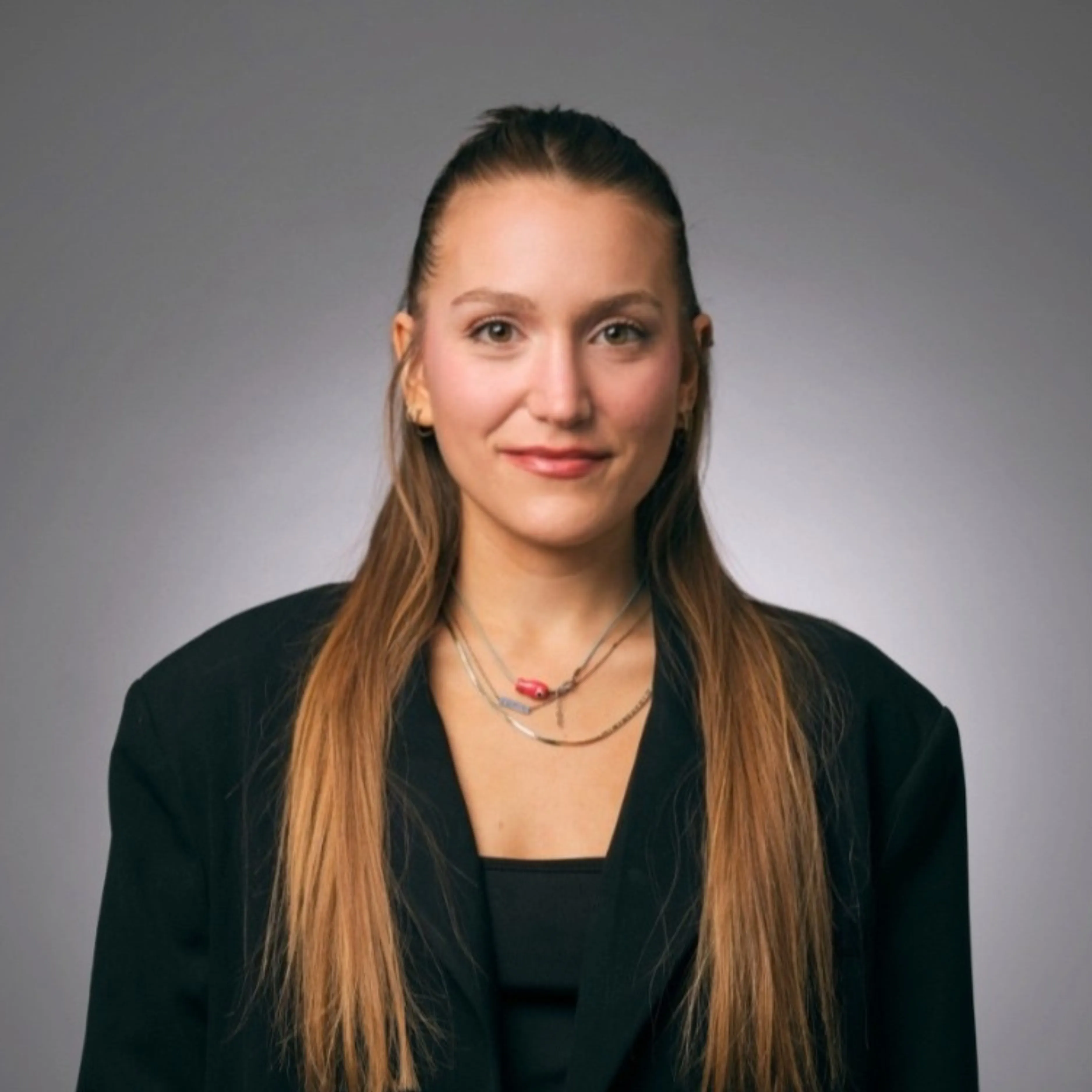 Young woman with long straight hair wearing a black blazer and layered necklaces against gray background.