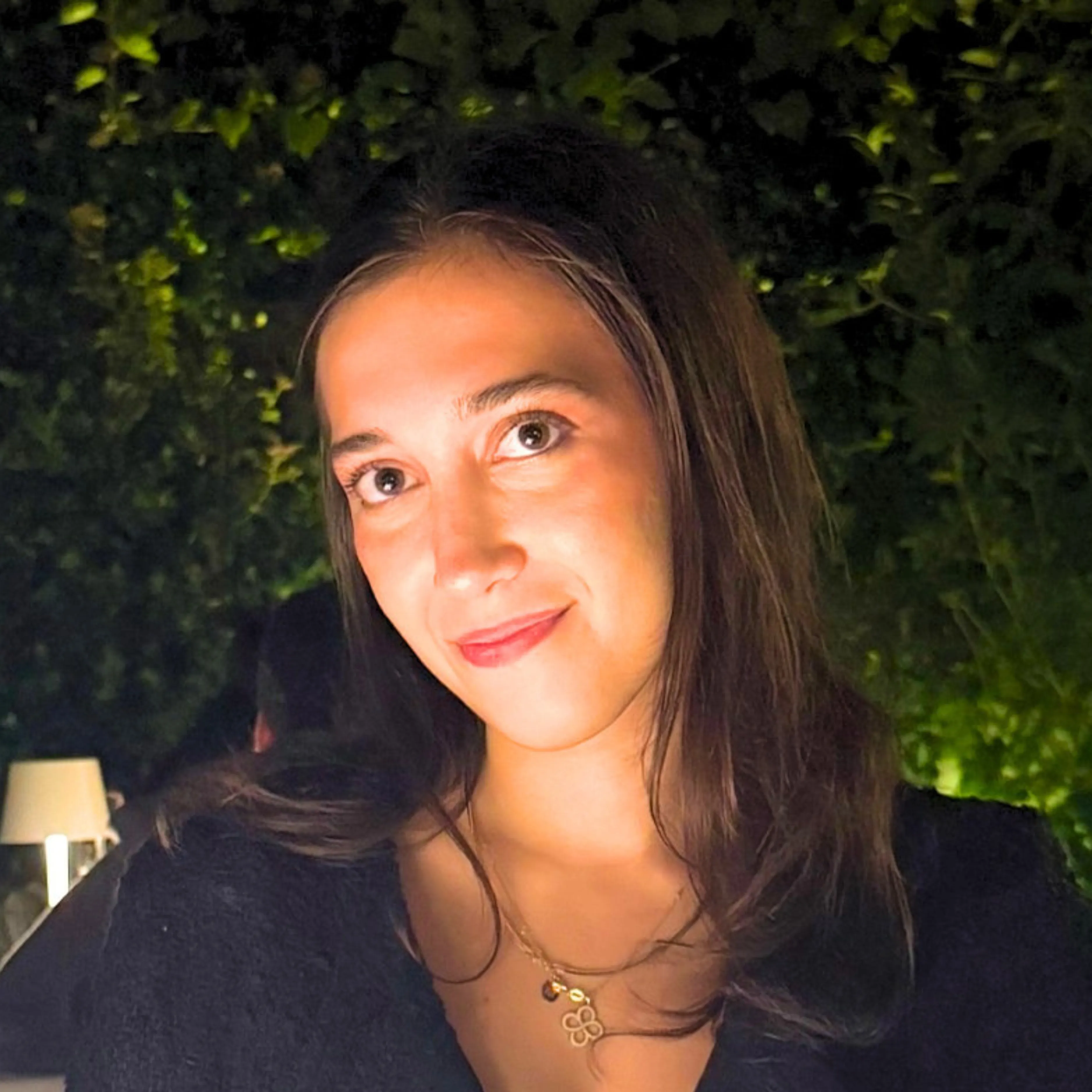 Young woman with long brown hair, wearing a black top and a gold necklace, smiling softly at night.