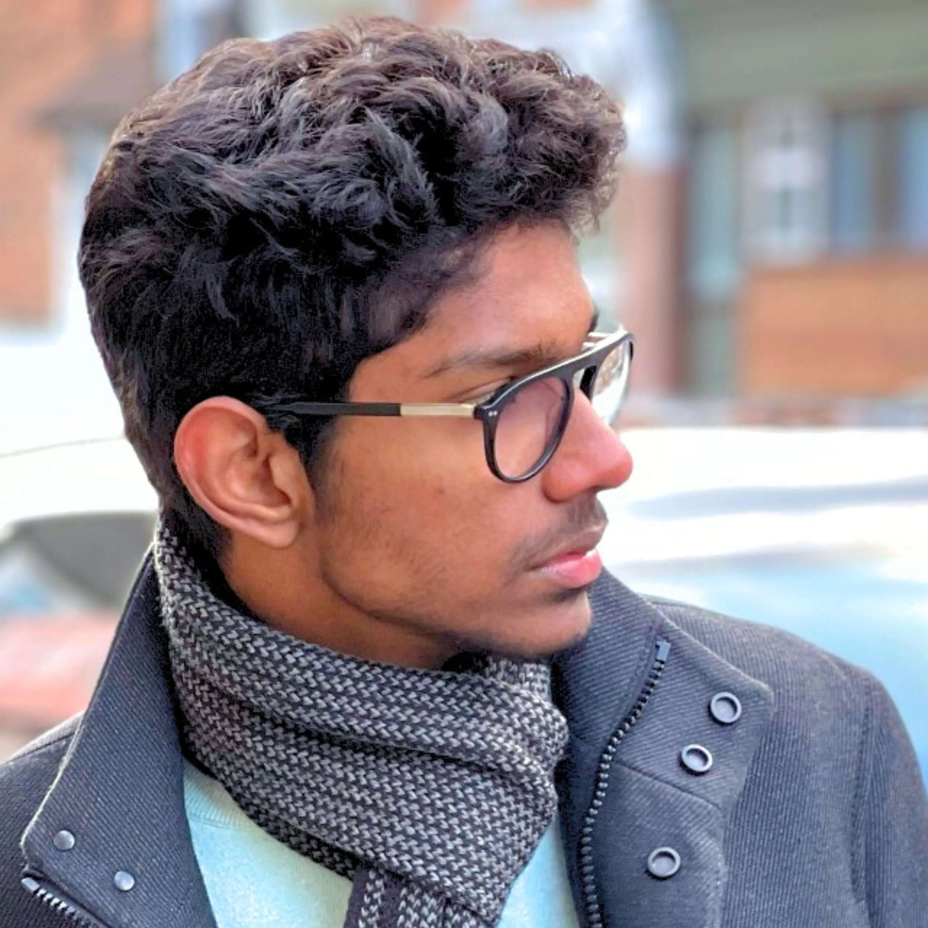 Young man with curly hair and black glasses wearing a gray scarf and jacket looking to the right.