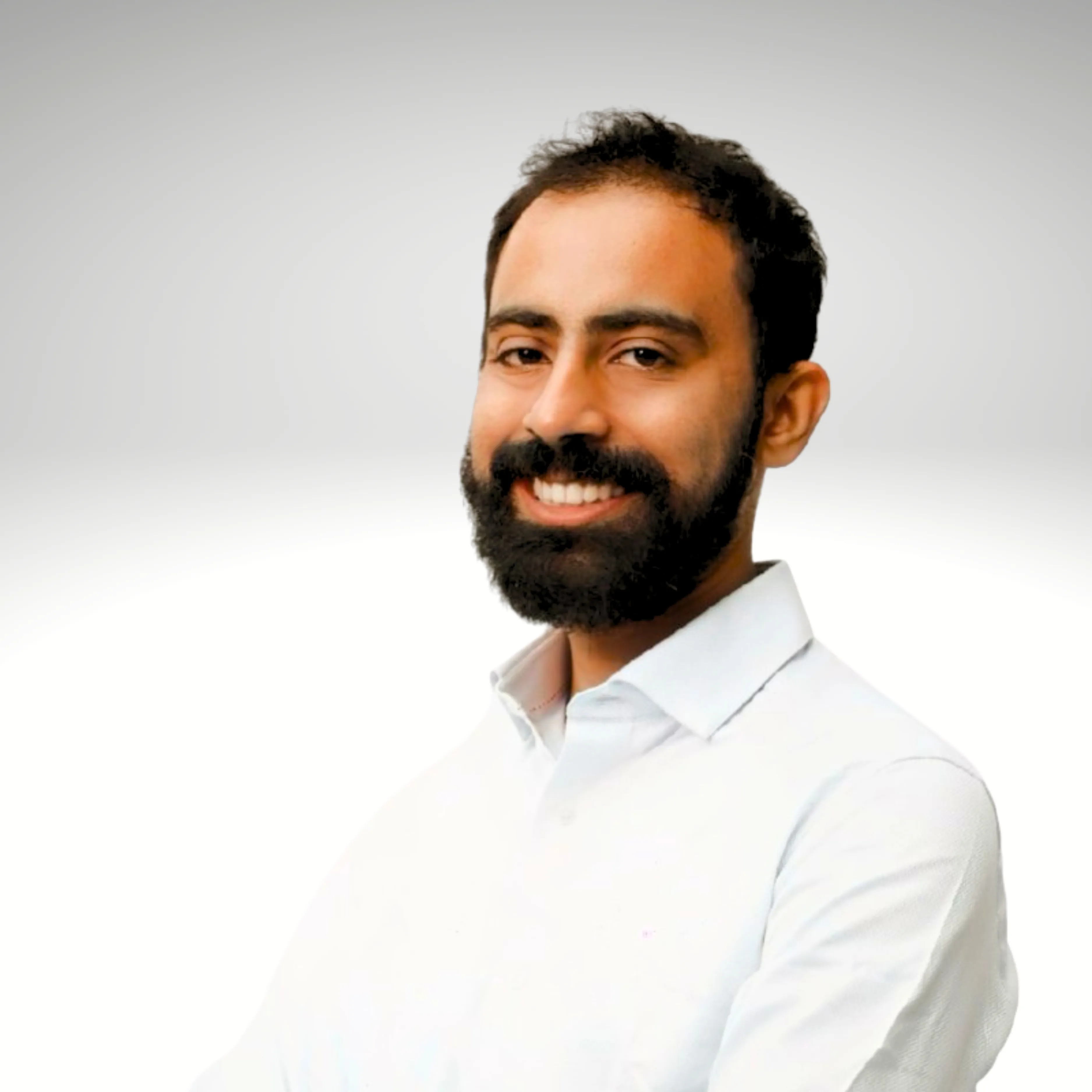 Smiling bearded man with dark hair wearing a white shirt against a gradient white background.