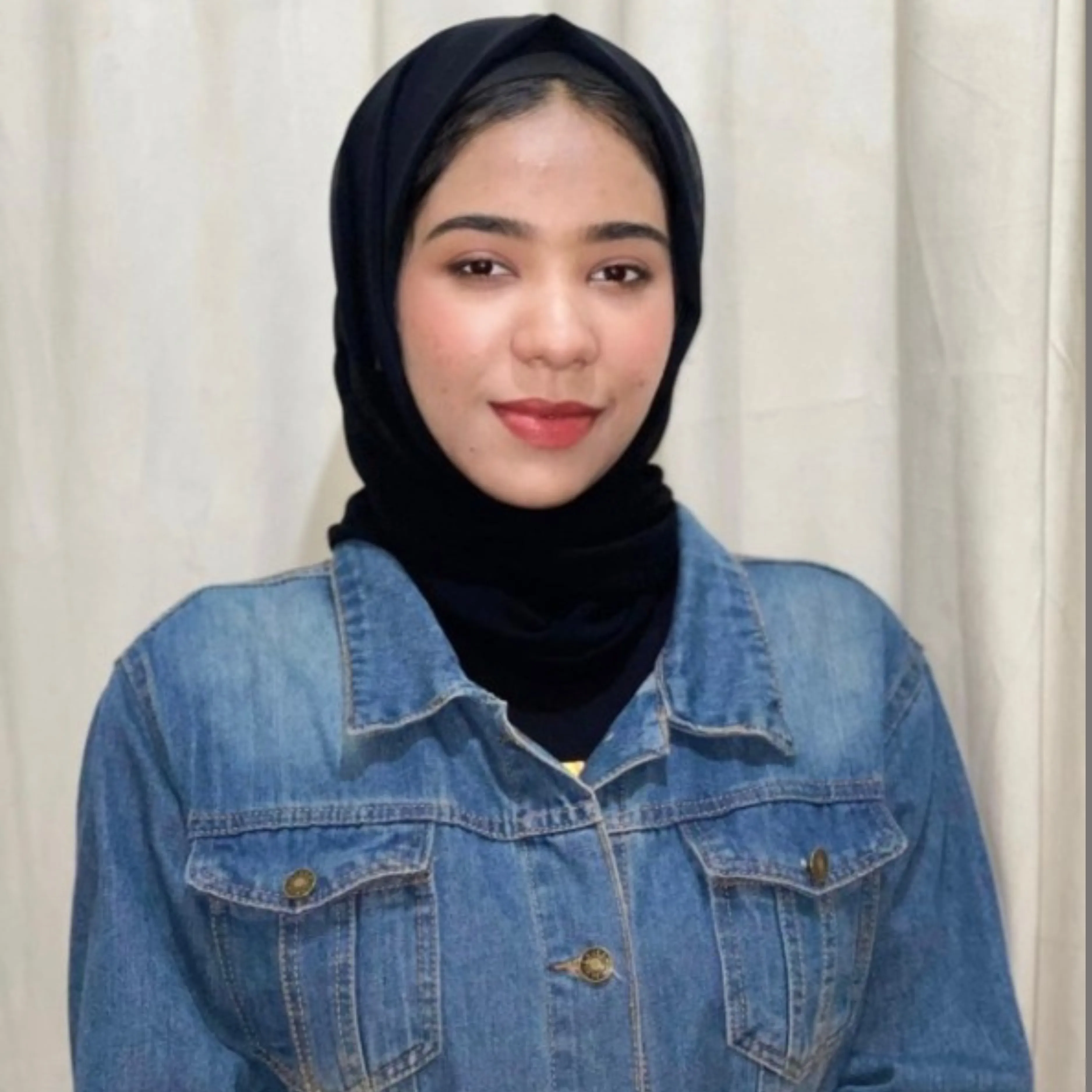 Young woman wearing a black hijab and denim jacket smiling softly against a light background.