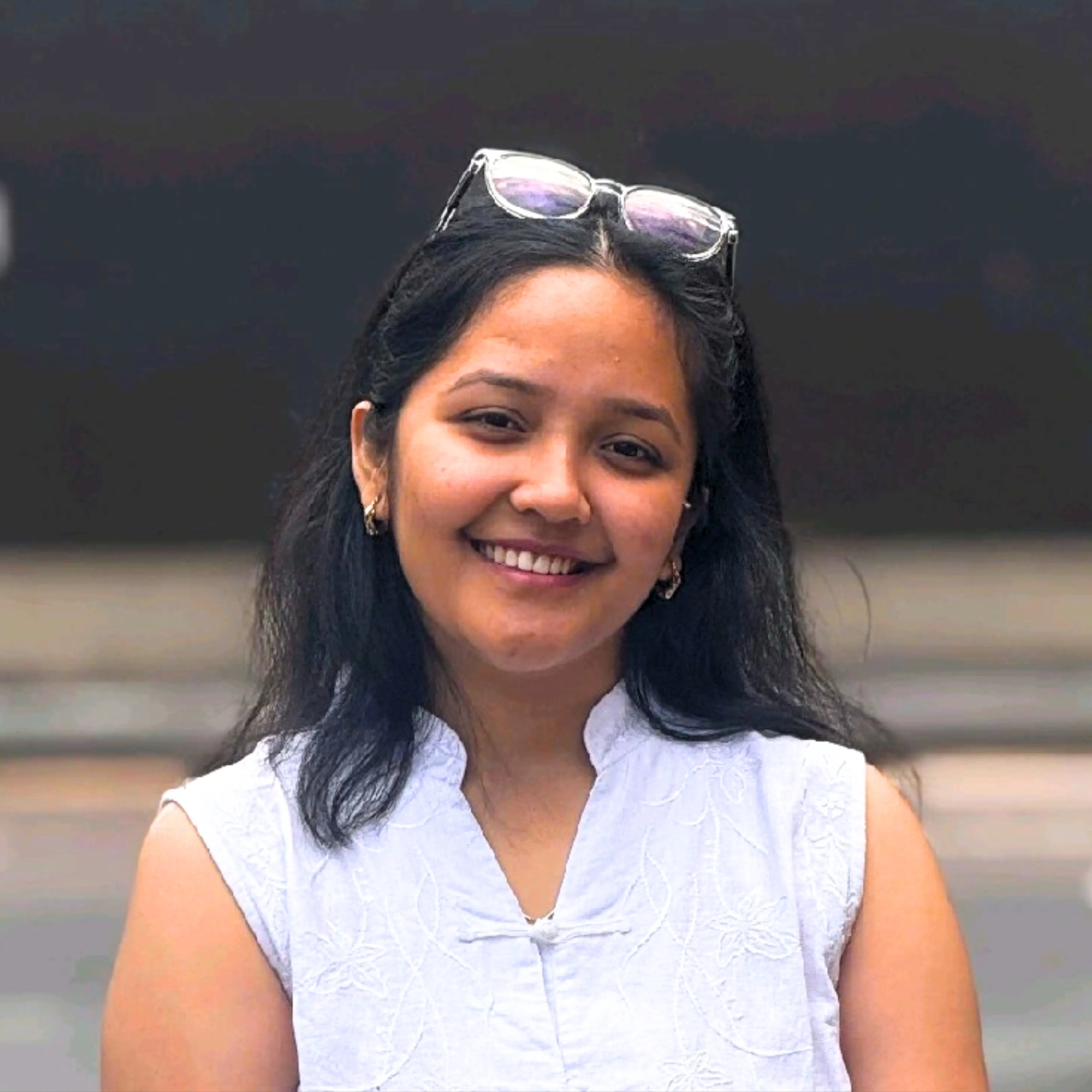 Young woman with long black hair smiling, wearing a white sleeveless top and sunglasses on her head.