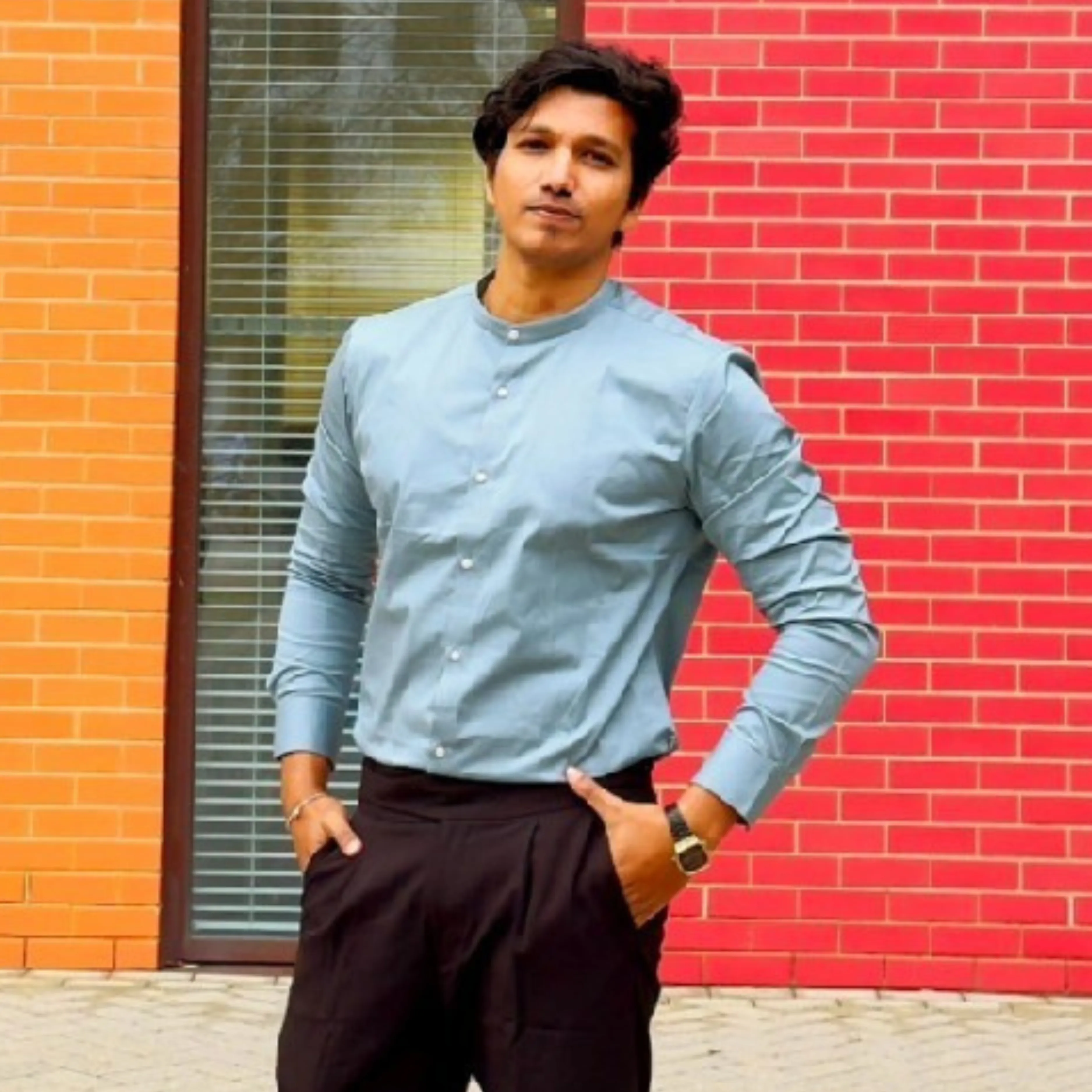 Man in light blue shirt and black pants standing with hands in pockets against brick wall.