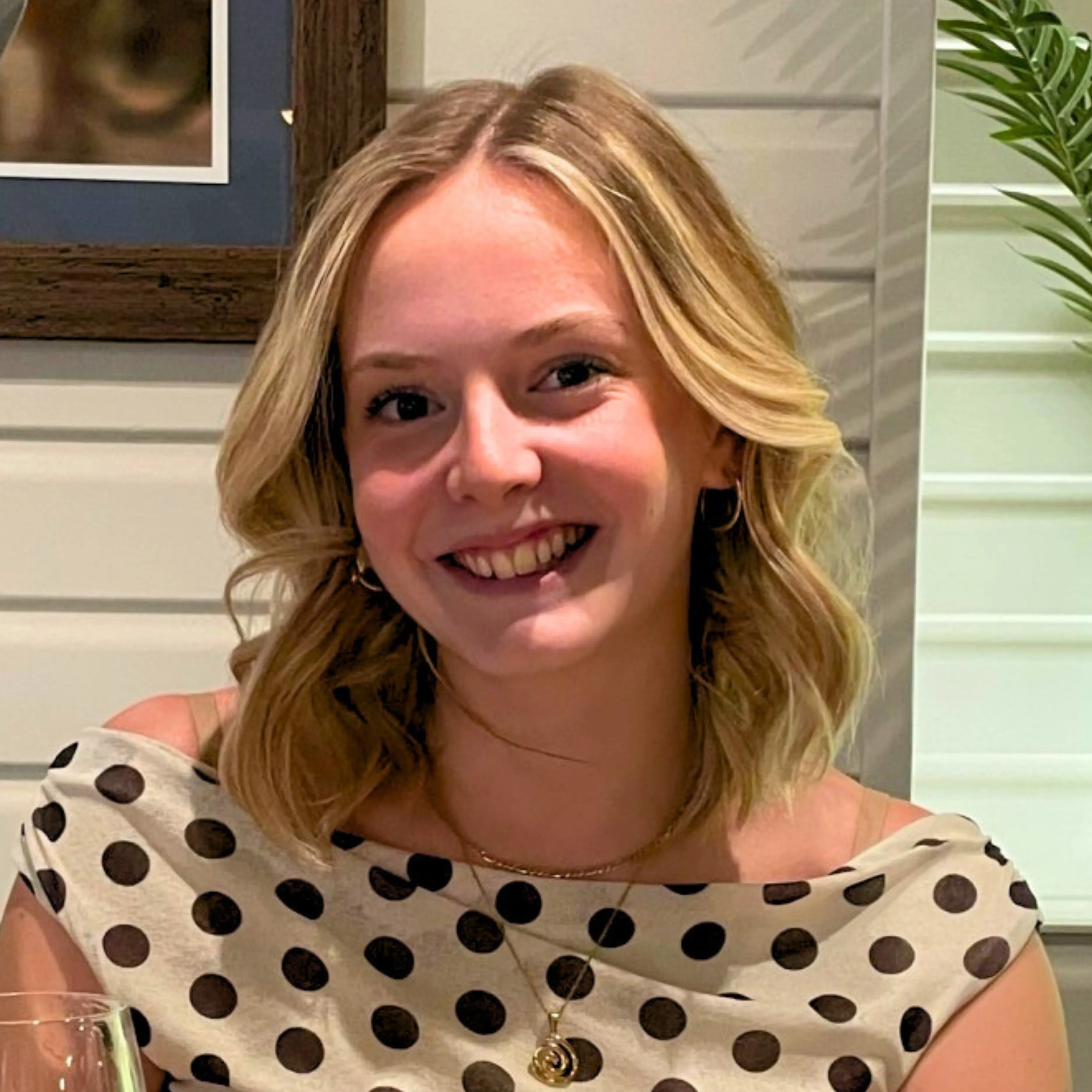 Smiling blonde woman with wavy hair wearing a polka dot top and gold necklace indoors.