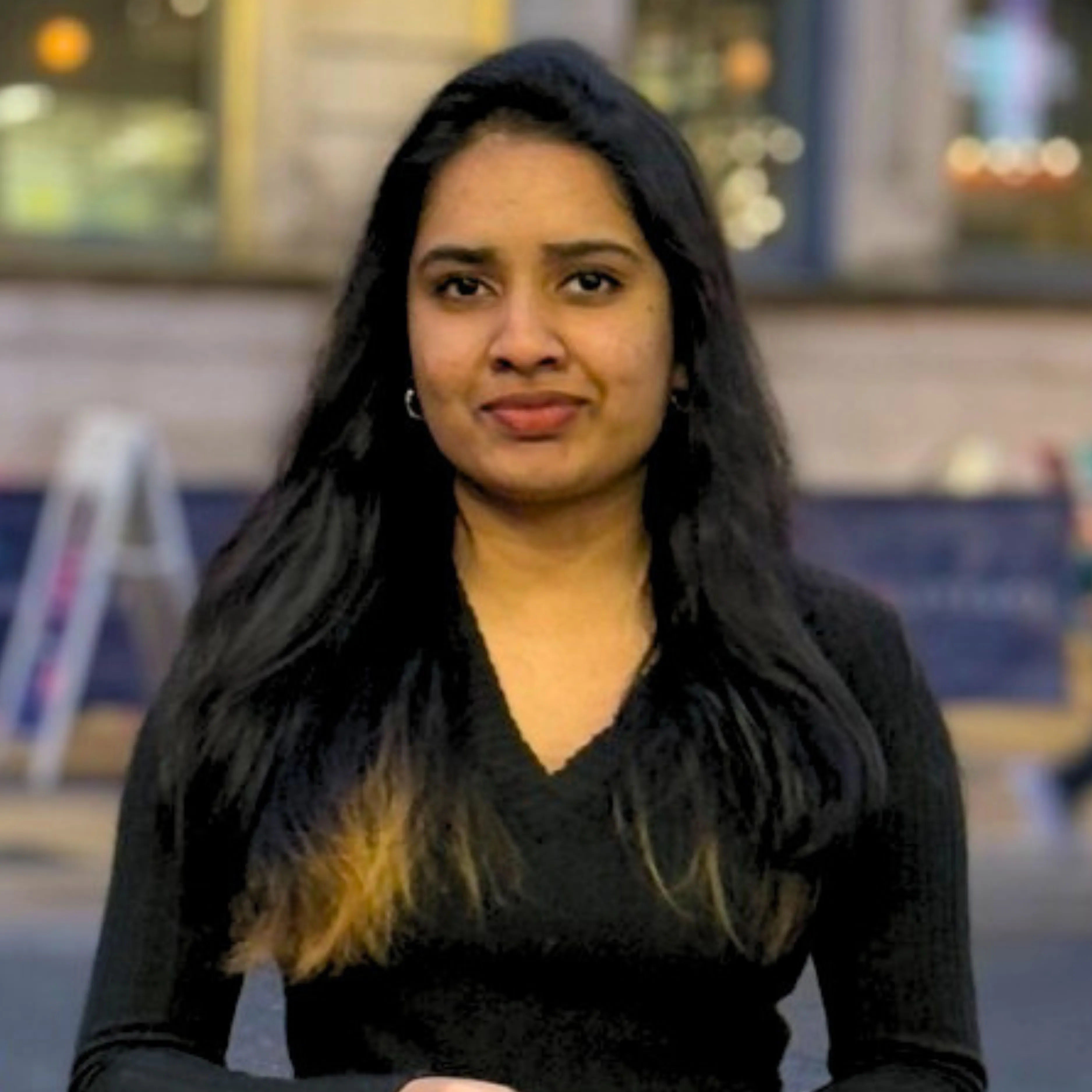Woman with long dark hair wearing a black sweater, standing outdoors with blurred background.