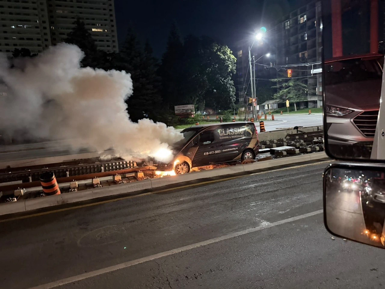 Vehicle fire beside a roadway at night with emergency responders nearby.
