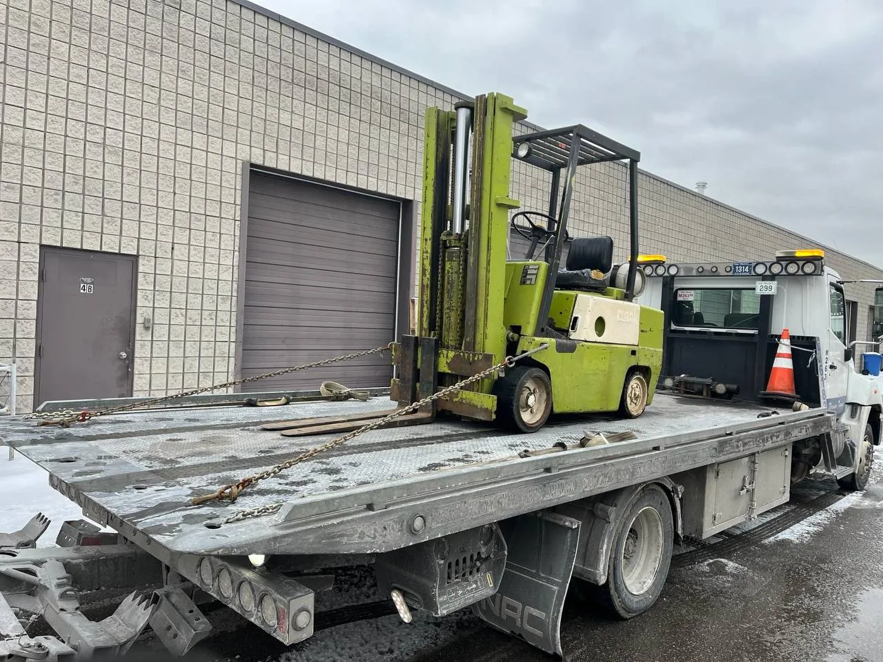 Green forklift secured on a tilt-and-load flatbed outside an industrial building.
