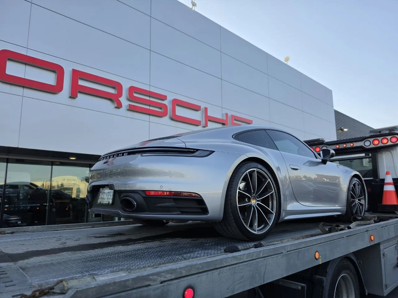 Silver Porsche convertible on a flatbed outside a Porsche dealership.