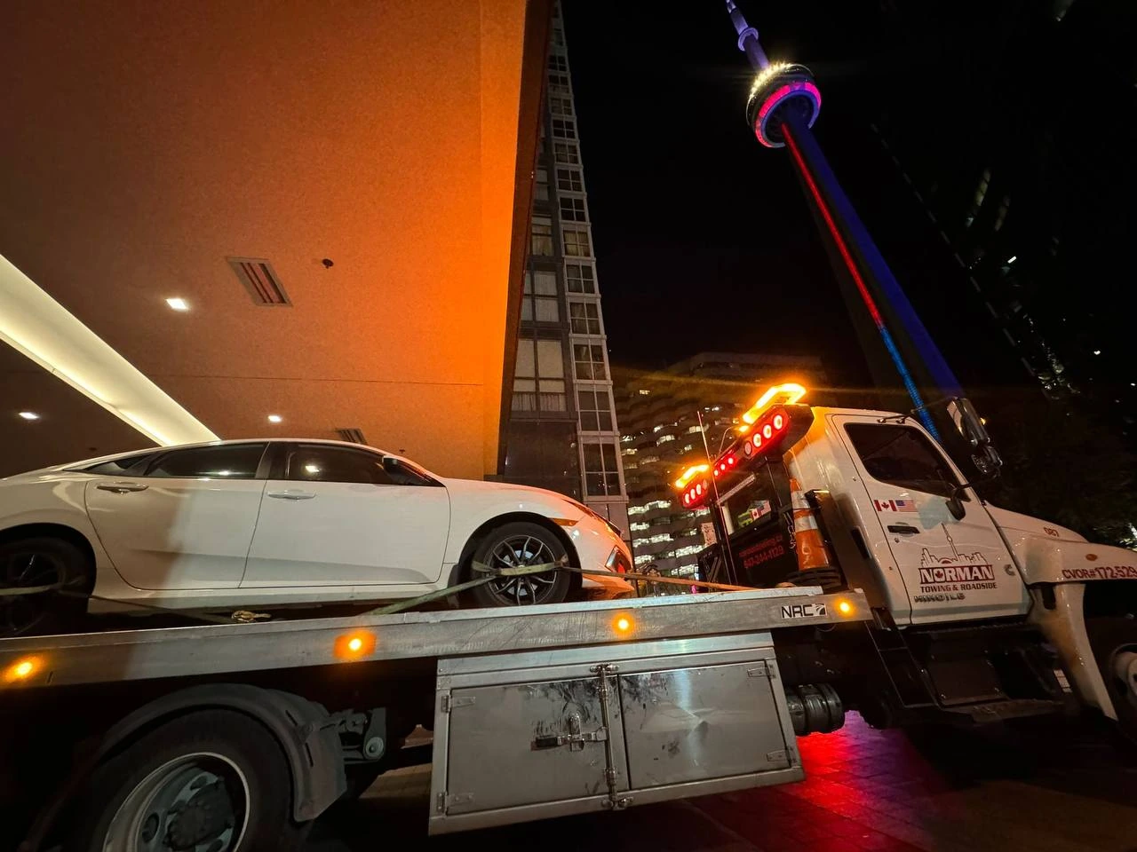White car on a Norman Towing flatbed near the CN Tower at night.