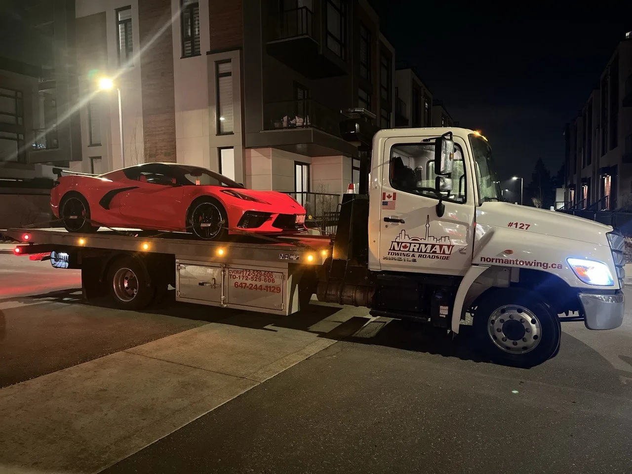 Red Corvette on a Norman Towing flatbed outside a residential building at night.