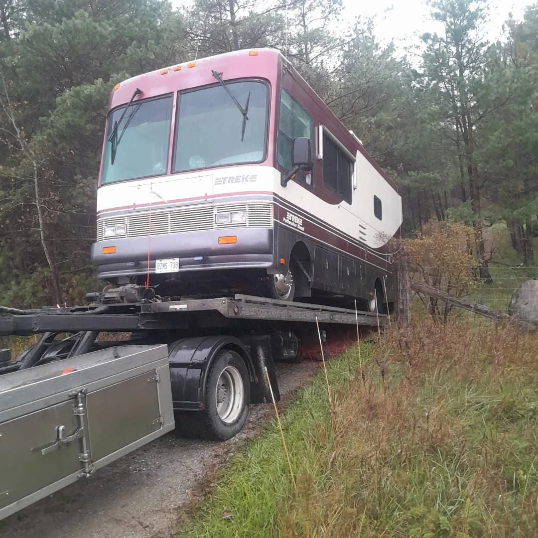 Older motorhome being loaded onto a flatbed in a wooded area.