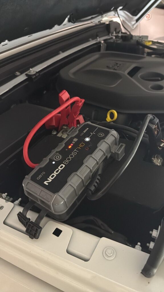 Close-up of a NOCO GB70 lithium jump starter hooked up to the passenger-side battery terminals of a Jeep Gladiator JT engine bay.