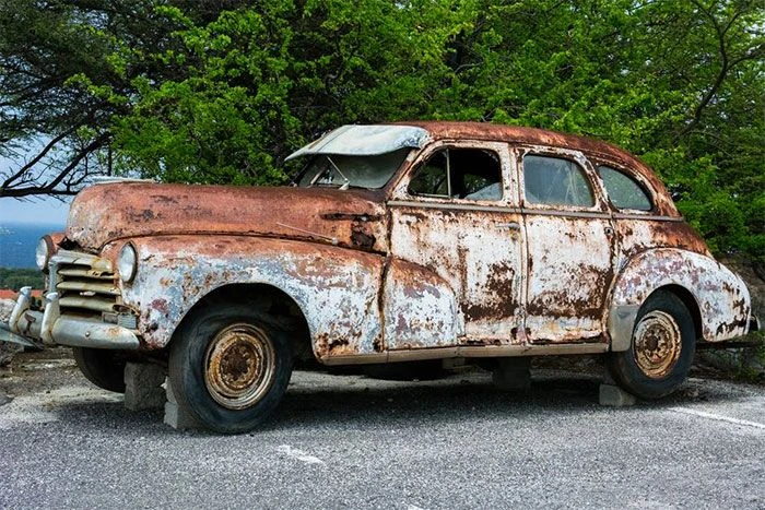 Rust-covered vintage car parked beside trees.