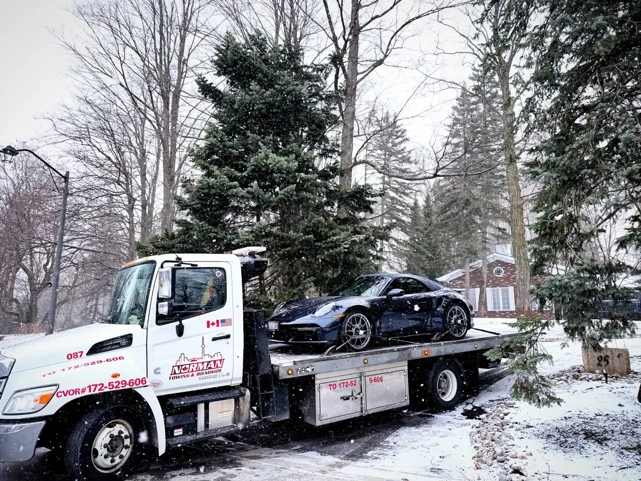 Norman Towing Flatbed In Falling Snow Norman Towing flatbed carrying a dark sports car on a snow-covered driveway during snowfall.