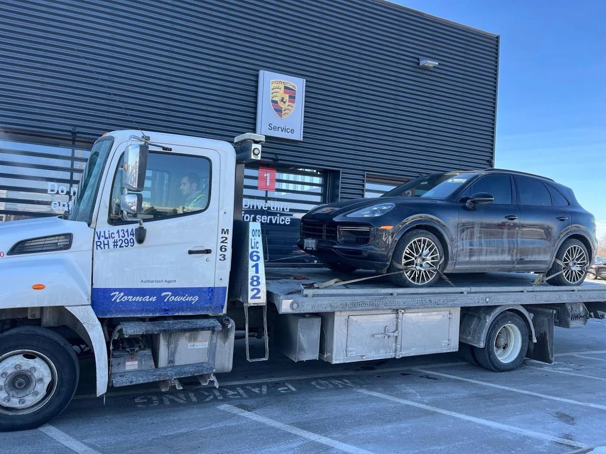 White Norman Towing flatbed carrying a dark Porsche SUV outside a Porsche service centre.