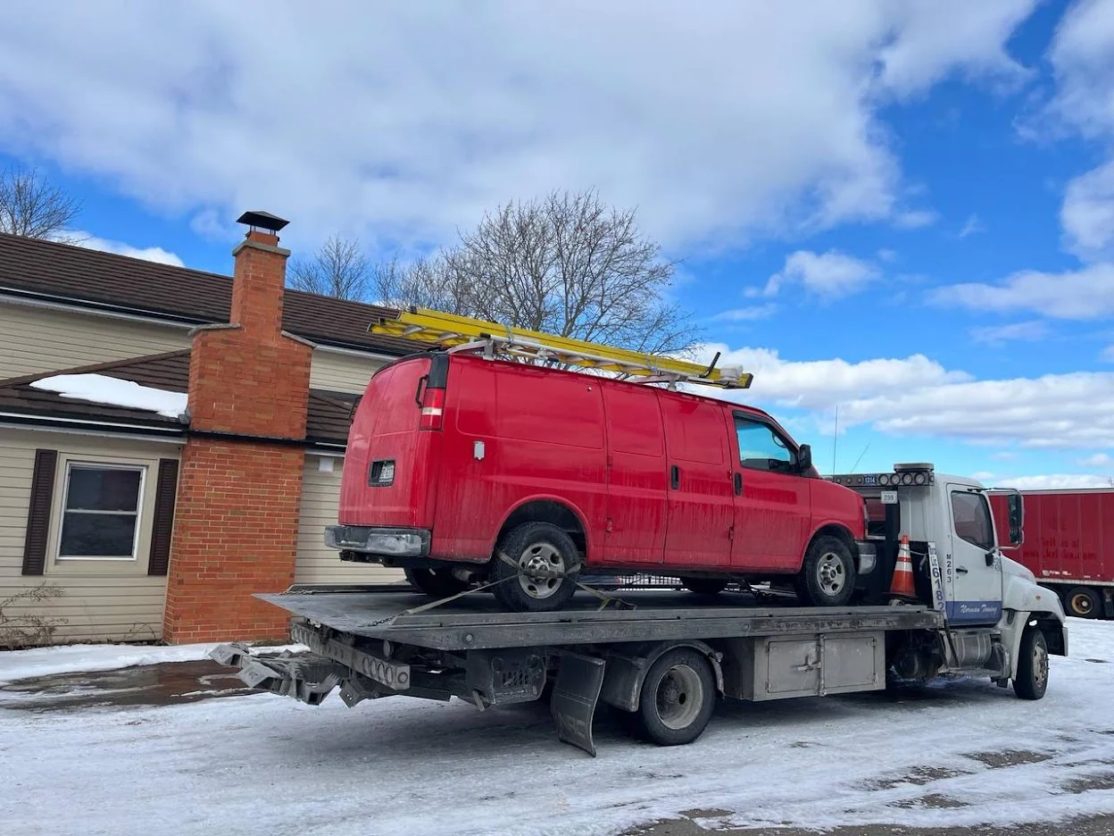 Norman Towing Flatbed Transporting A Red Work Van Red work van with roof ladders secured on a Norman Towing flatbed beside a snow-covered driveway.