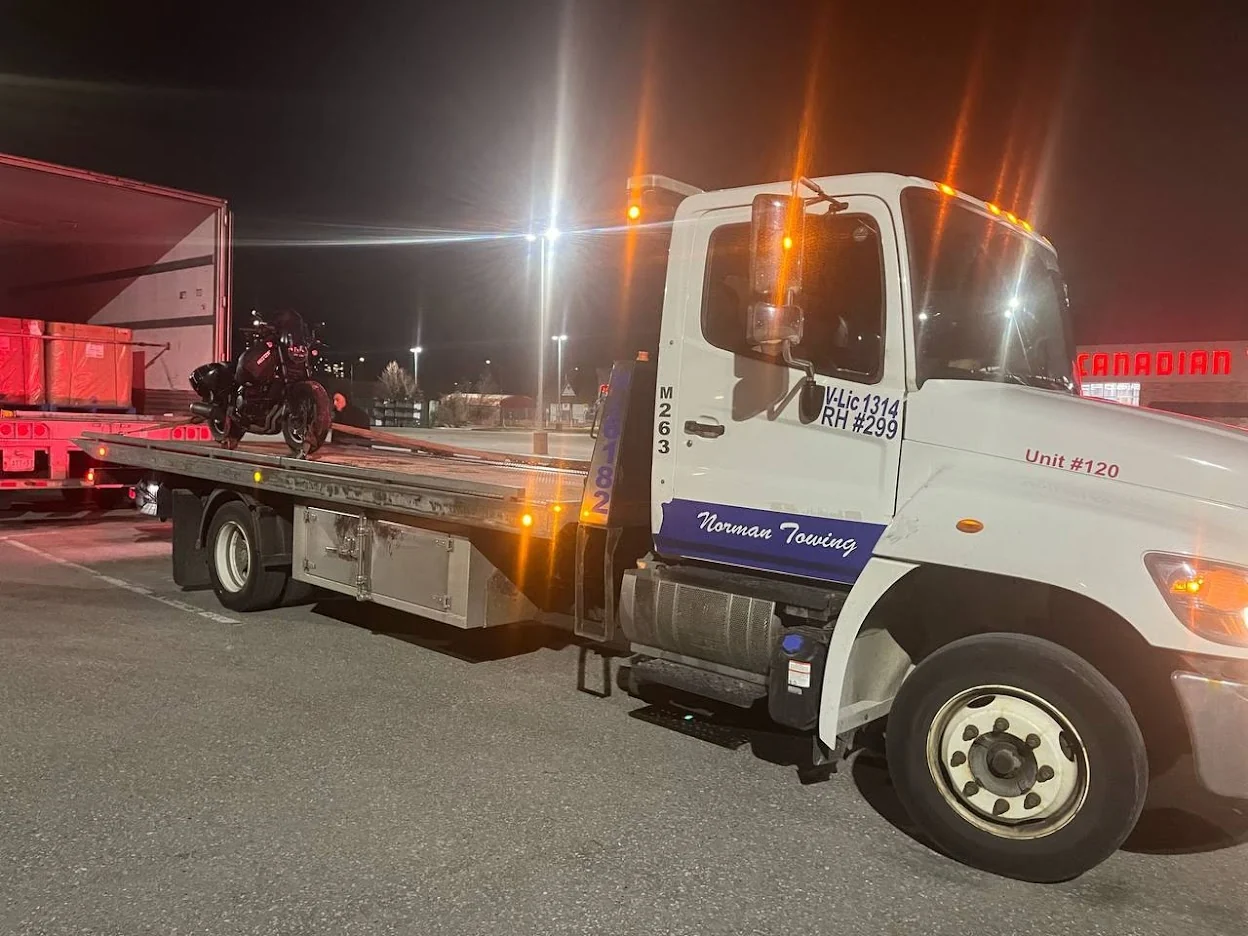 Norman Towing Motorcycle Transport At Night White Norman Towing flatbed carrying a motorcycle at night with amber warning lights illuminated.