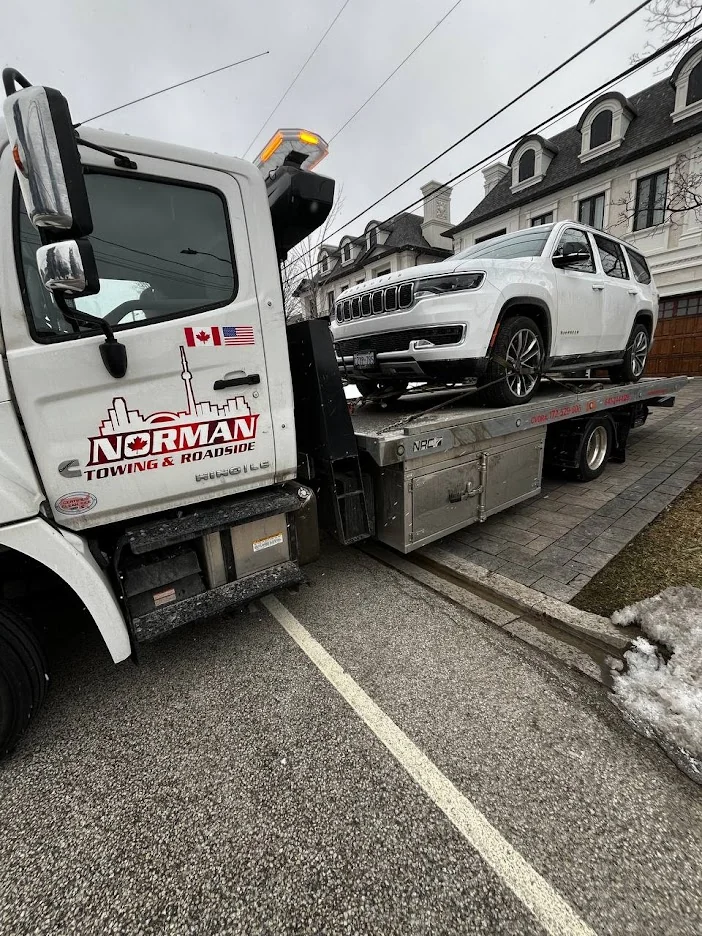 Norman Towing Flatbed Carrying A White Jeep White Jeep SUV secured on a Norman Towing flatbed beside a residential driveway.