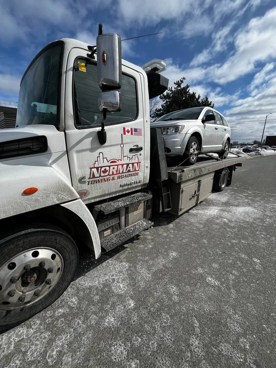 White flatbed truck with the red Norman Towing logo carrying a silver SUV beside patches of snow.