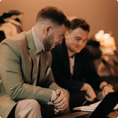 Two men in blazers smiling and looking at a laptop in a warmly lit room.