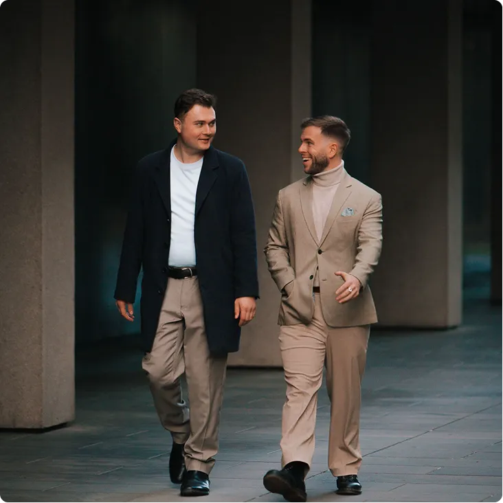 Two men dressed in smart casual attire walking and chatting outdoors near concrete pillars.