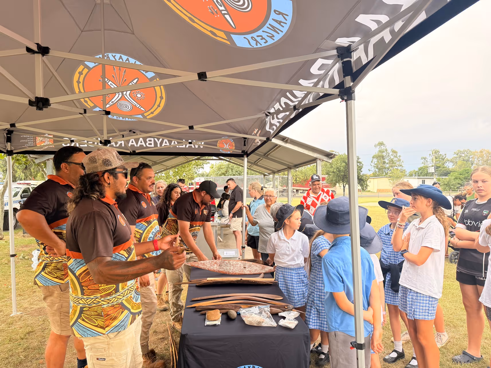 Group of Walaaybaa Rangers showing Aboriginal artifacts and art to schoolchildren under a tent outdoors.