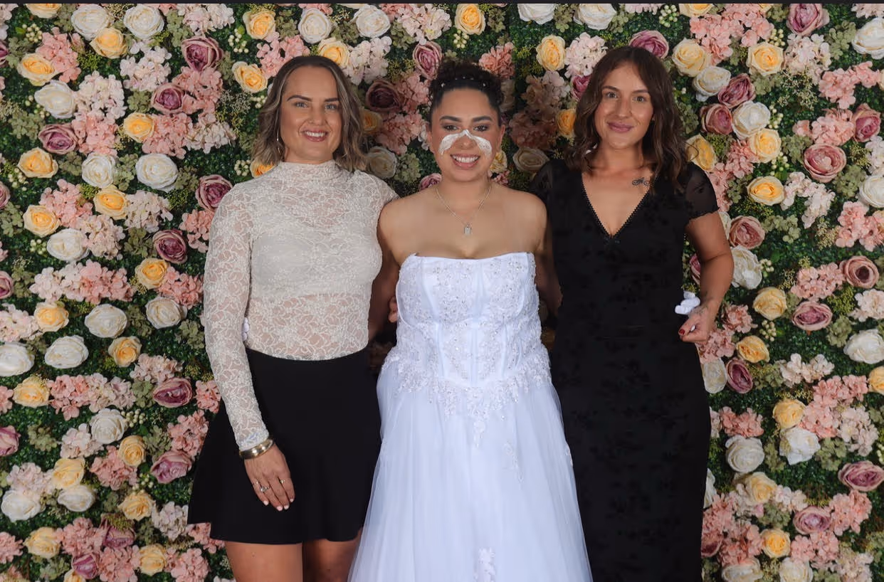 3 women stand in front of a floral background, the woman in the middle wears a white dress, and traditional ochre face paint as part of her NAIDOC debutante ball.