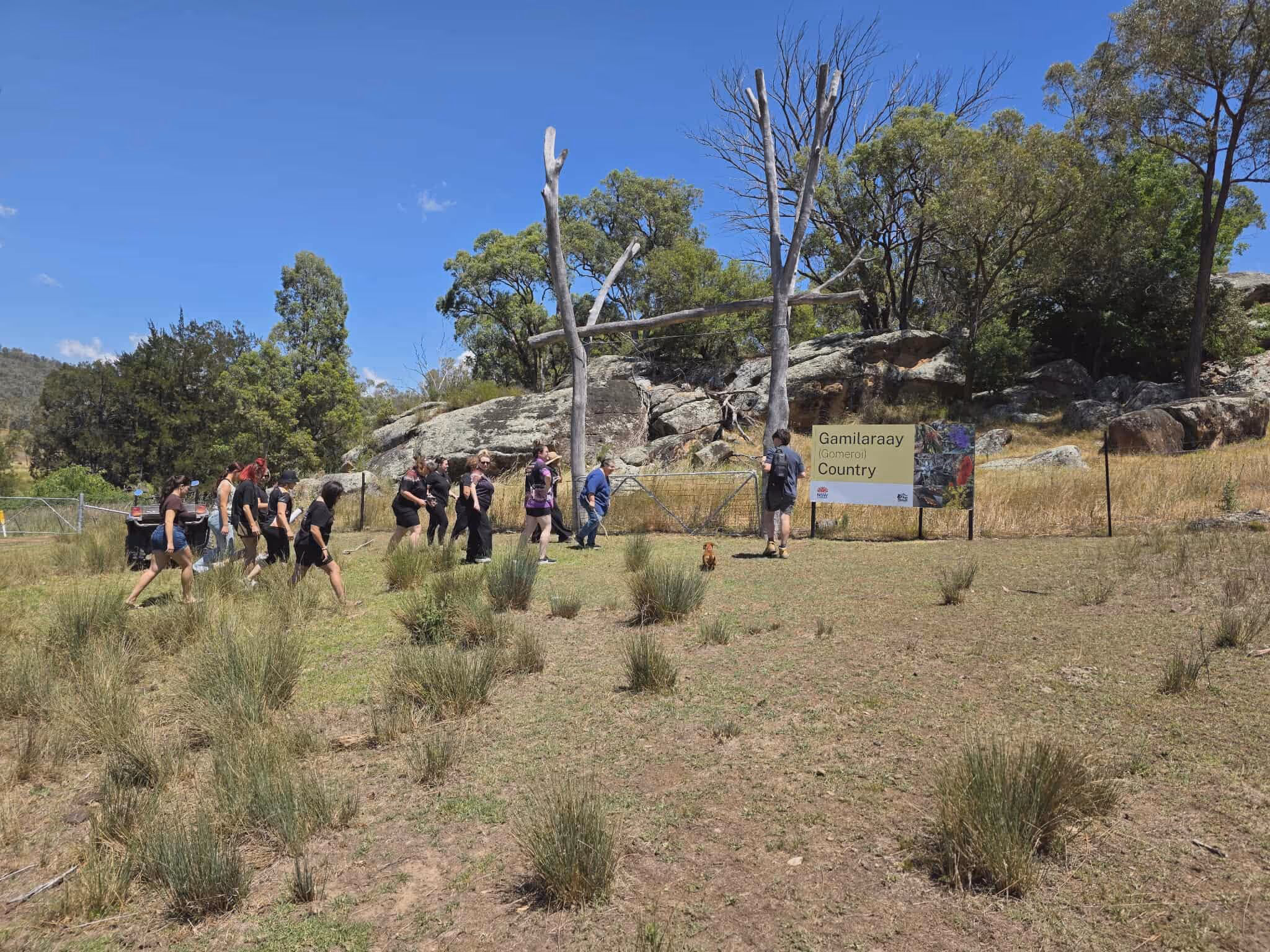 A group of people waking through the bush in front of an entry way made out of old trees