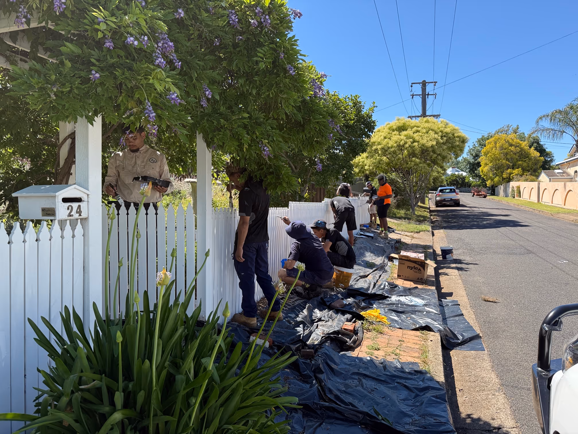 Fresh footprints participants are in a green front yard, painting a wood picket fence a fresh bright white