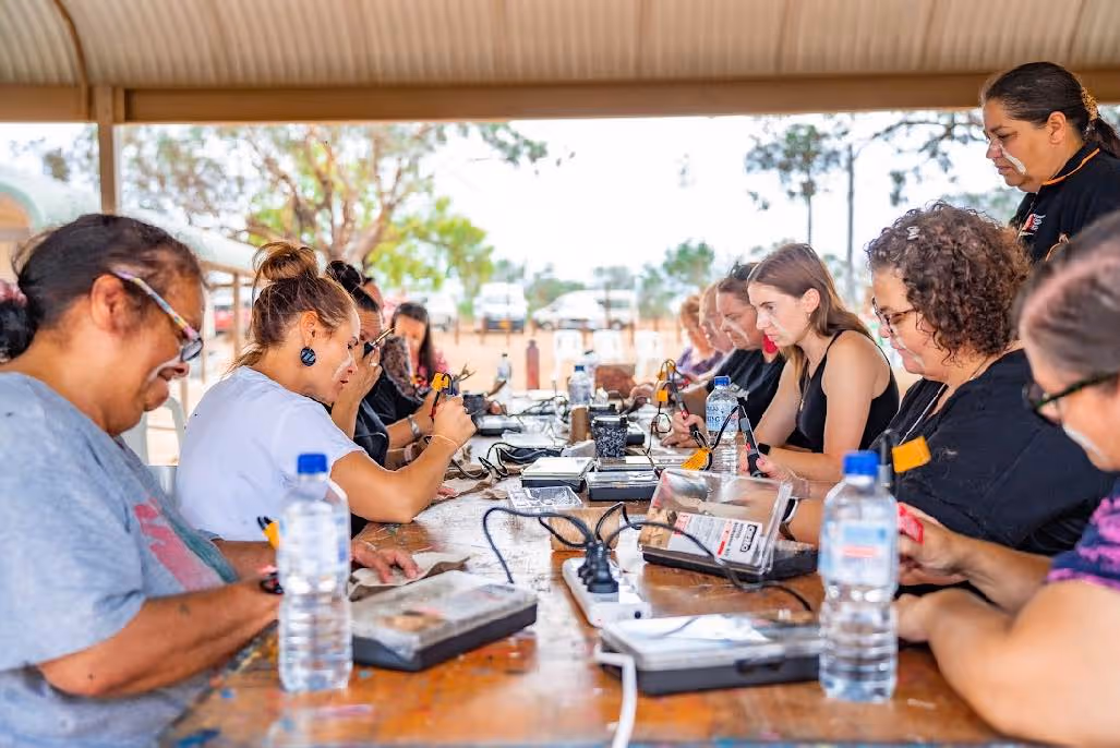 Group of women sitting at a long table using electronic devices with tools under a covered outdoor area.