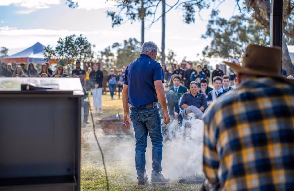 Len Waters wears blue shirt, performing a smoking ceremony in front of seated audience outdoors on a sunny day.