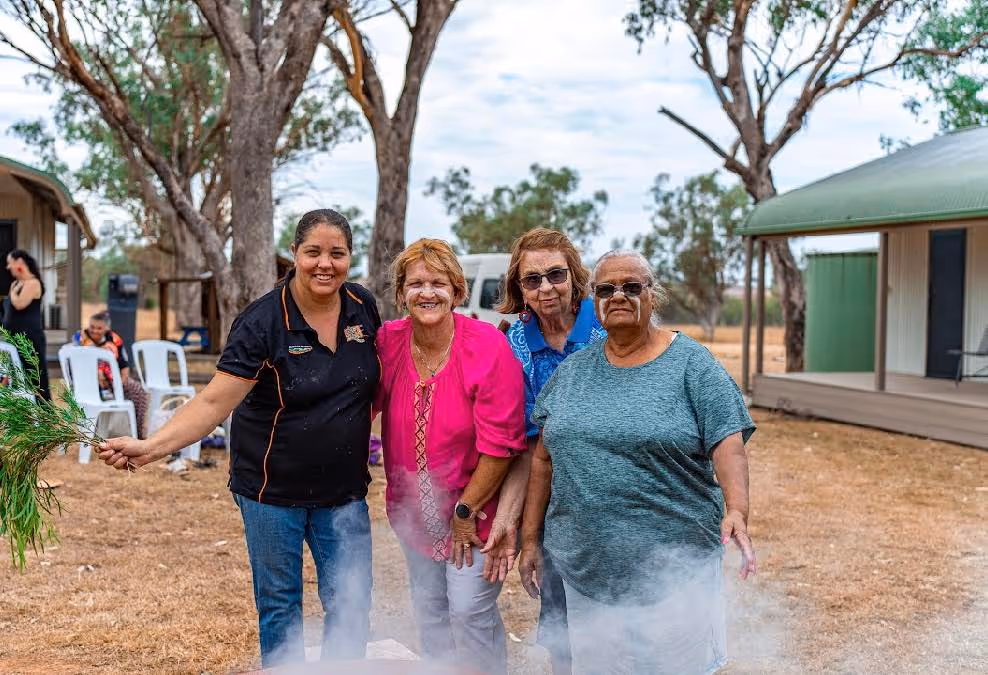 Four women standing outdoors smiling, one holding green leaves, with smoke rising in front of them.