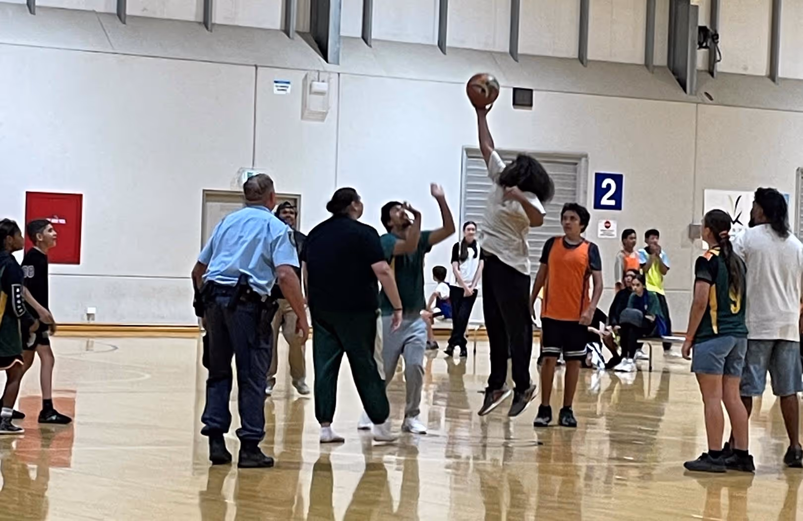 Bounce Back participants and guest police officers playing basketball indoors, one player jumping with ball extended to the hoop.
