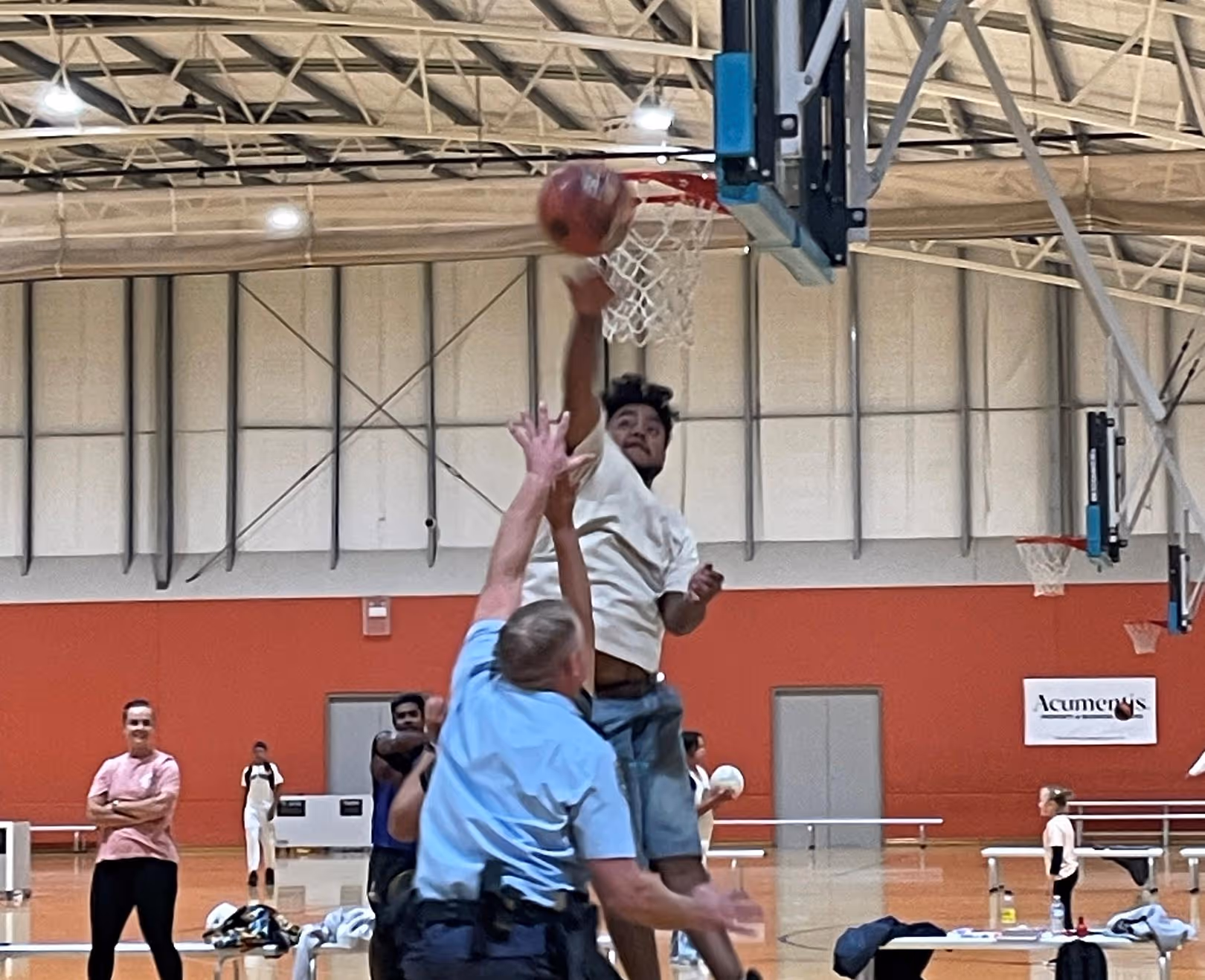 Bounce Back participants and community police officer jumping near basketball hoop inside gym with spectators seated and standing in background.