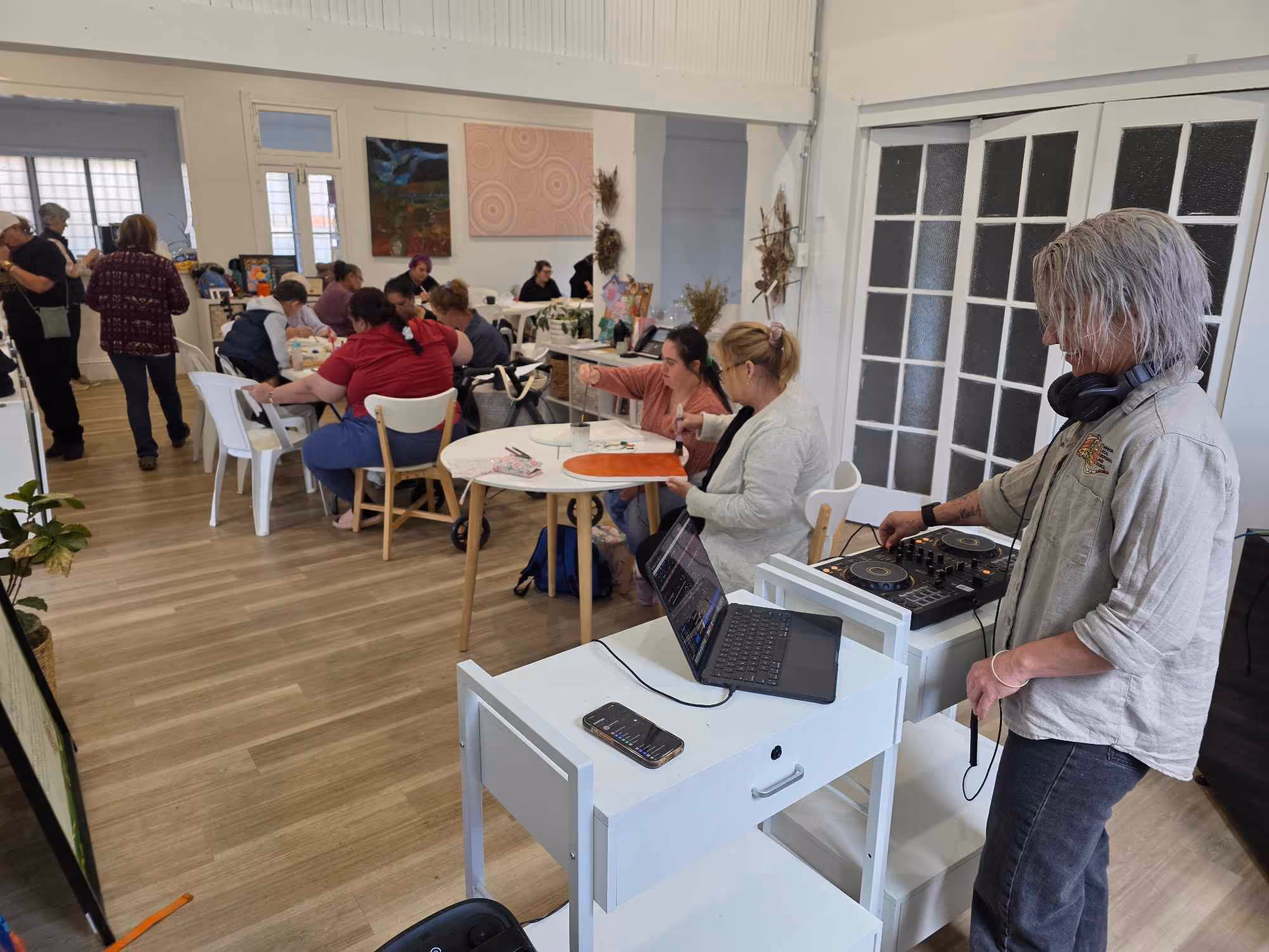 A group of people painting and socializing in a well-lit art studio; a DJ controls music in the foreground.