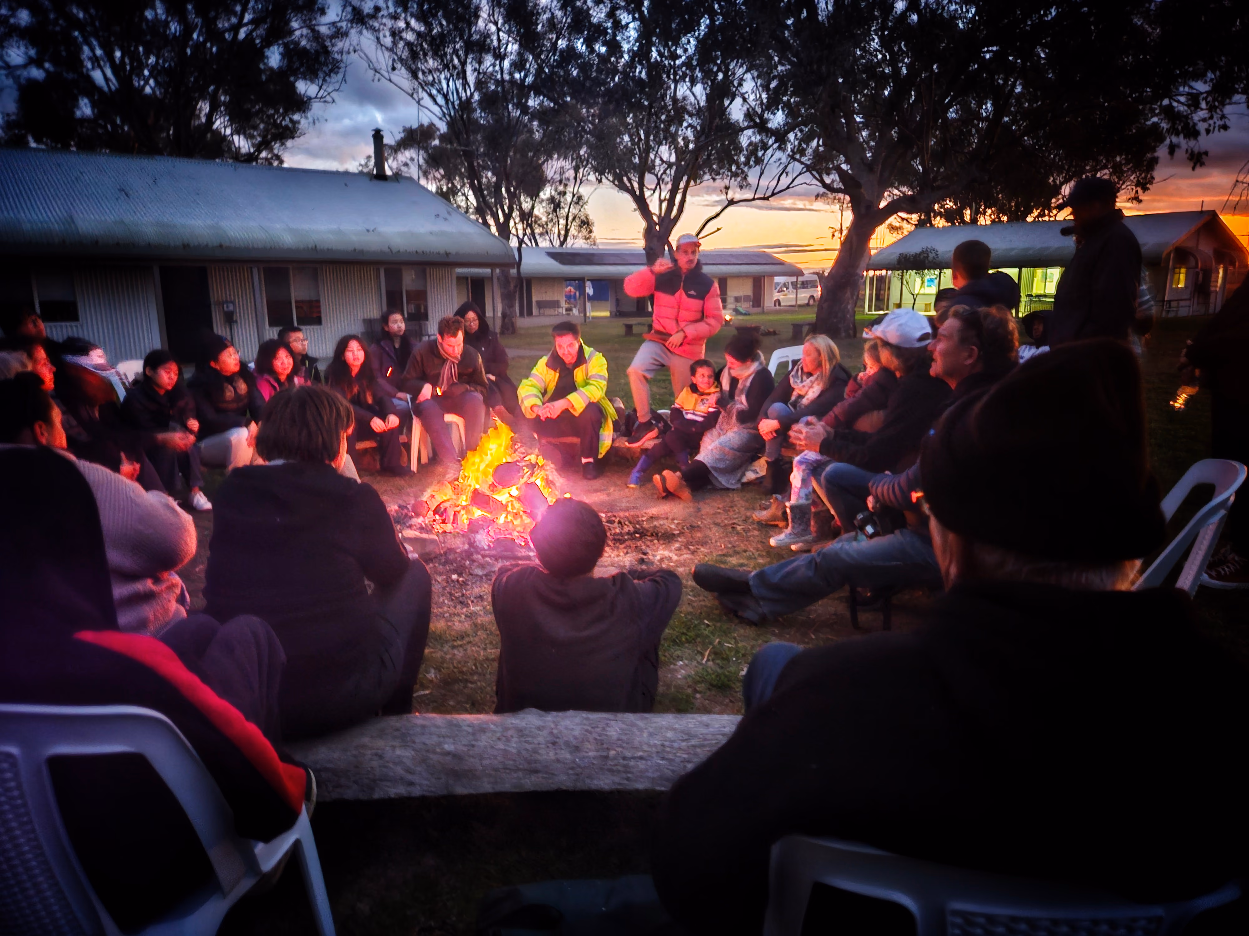 Group of people sitting around a campfire outdoors at dusk near cabins and trees.