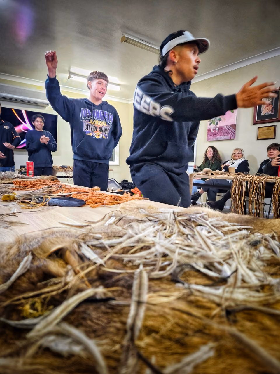 Young people in a room performing, with others seated watching, and hides with feathers on tables.